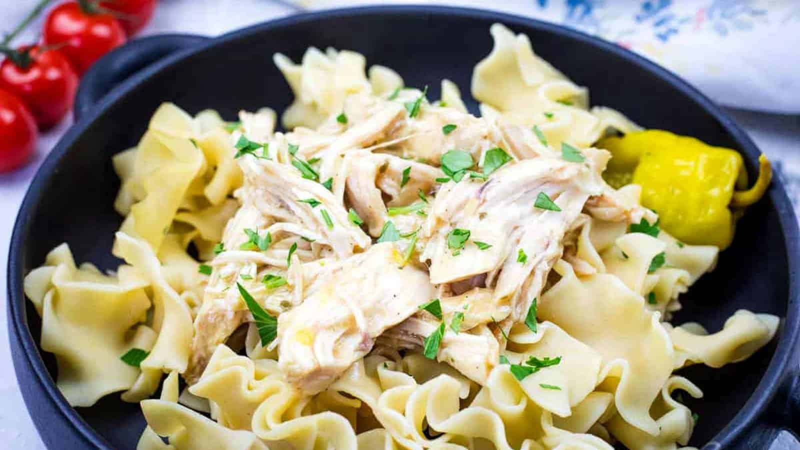 A black bowl containing egg noodles topped with Slow Cooker Mississippi Chicken and garnished with parsley, accompanied by a pickled pepper.