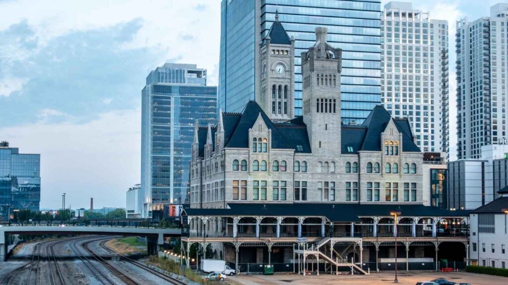 Historic stone building with clock tower surrounded by modern skyscrapers, adjacent to railway tracks under a cloudy sky.