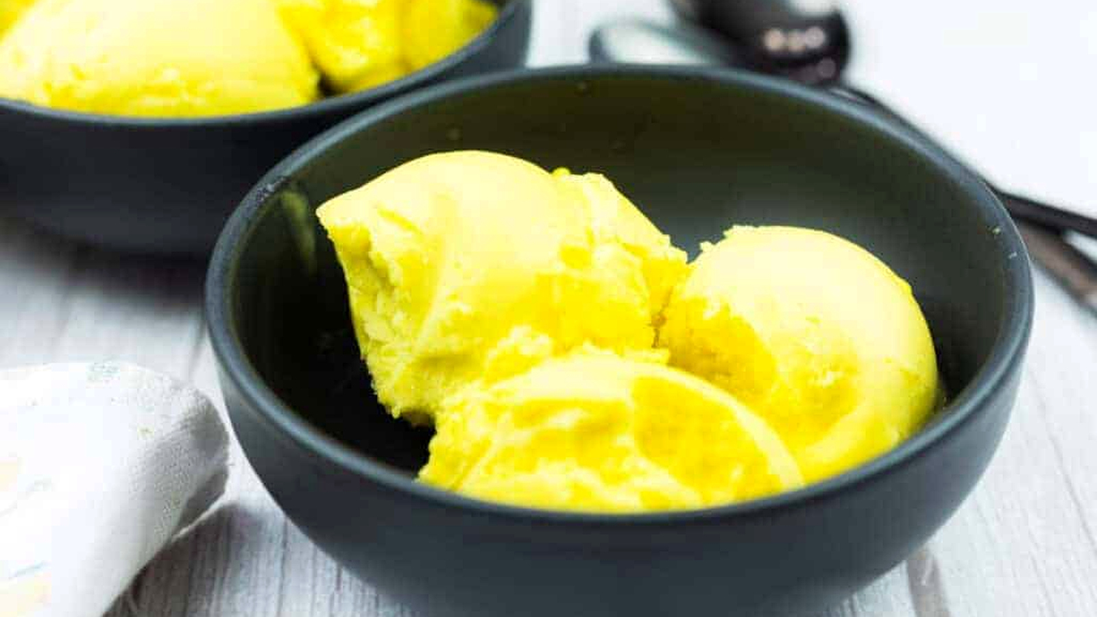 A black bowl containing three scoops of bright yellow ice cream sits on a light wooden surface, with another bowl and metal spoons in the background.