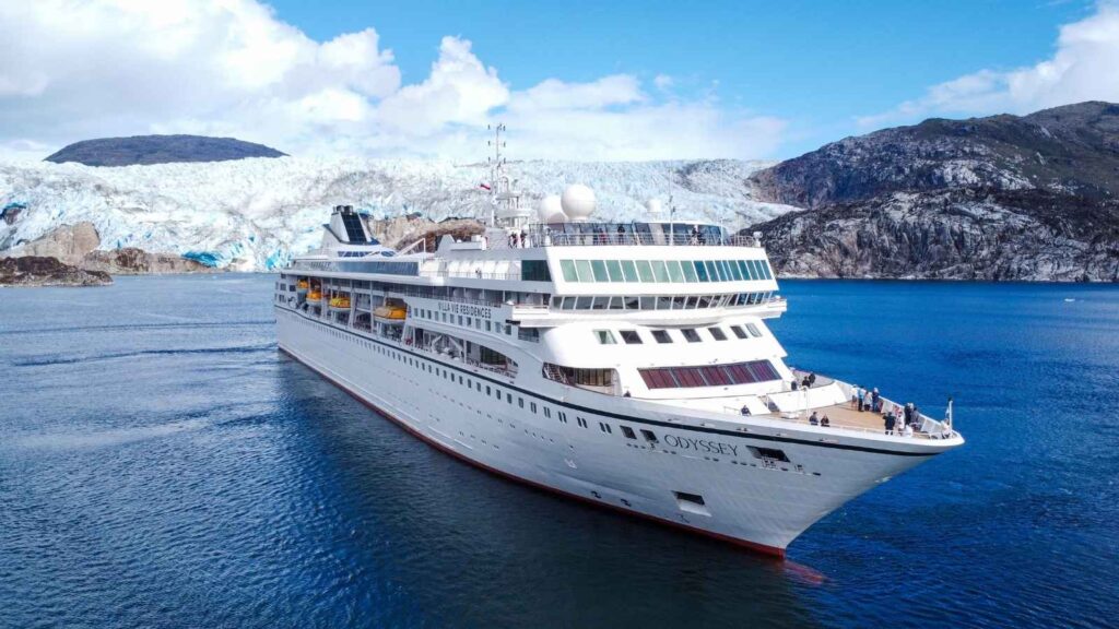 A white cruise ship named Odyssey sails on calm blue water with a glacier and rocky mountains in the background.
