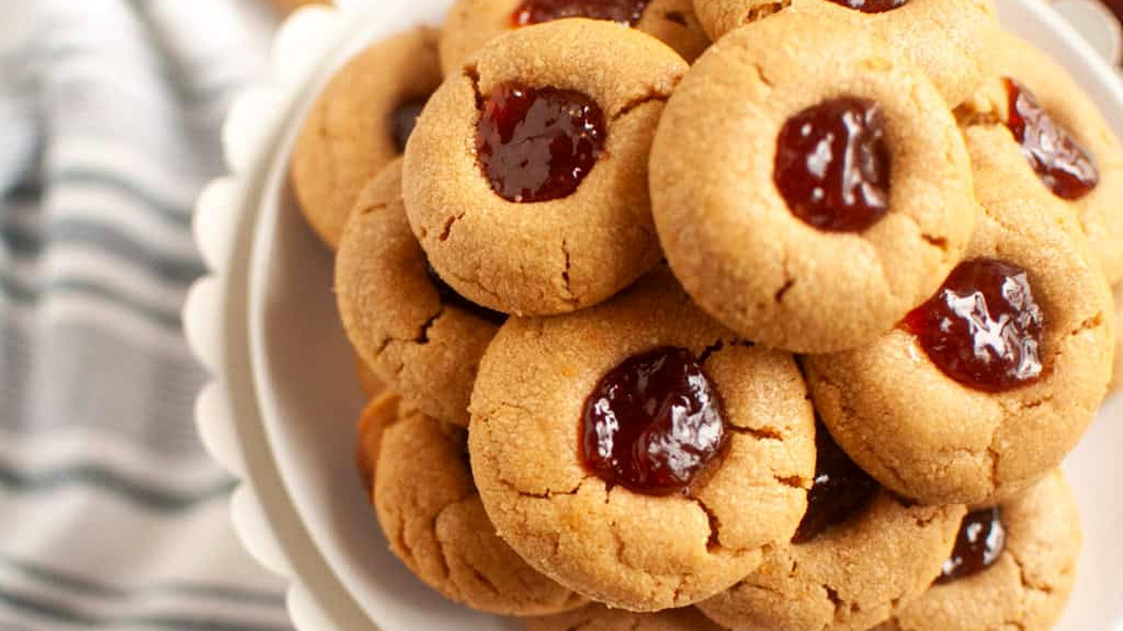 A plate stacked with Peanut butter jelly thumbprint cookies filled with red jam, placed on a white stand next to a striped cloth.