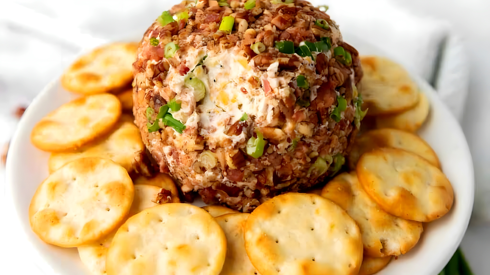 A hand holding a round cracker, reaching to dip into a pecan bacon cheeseball.