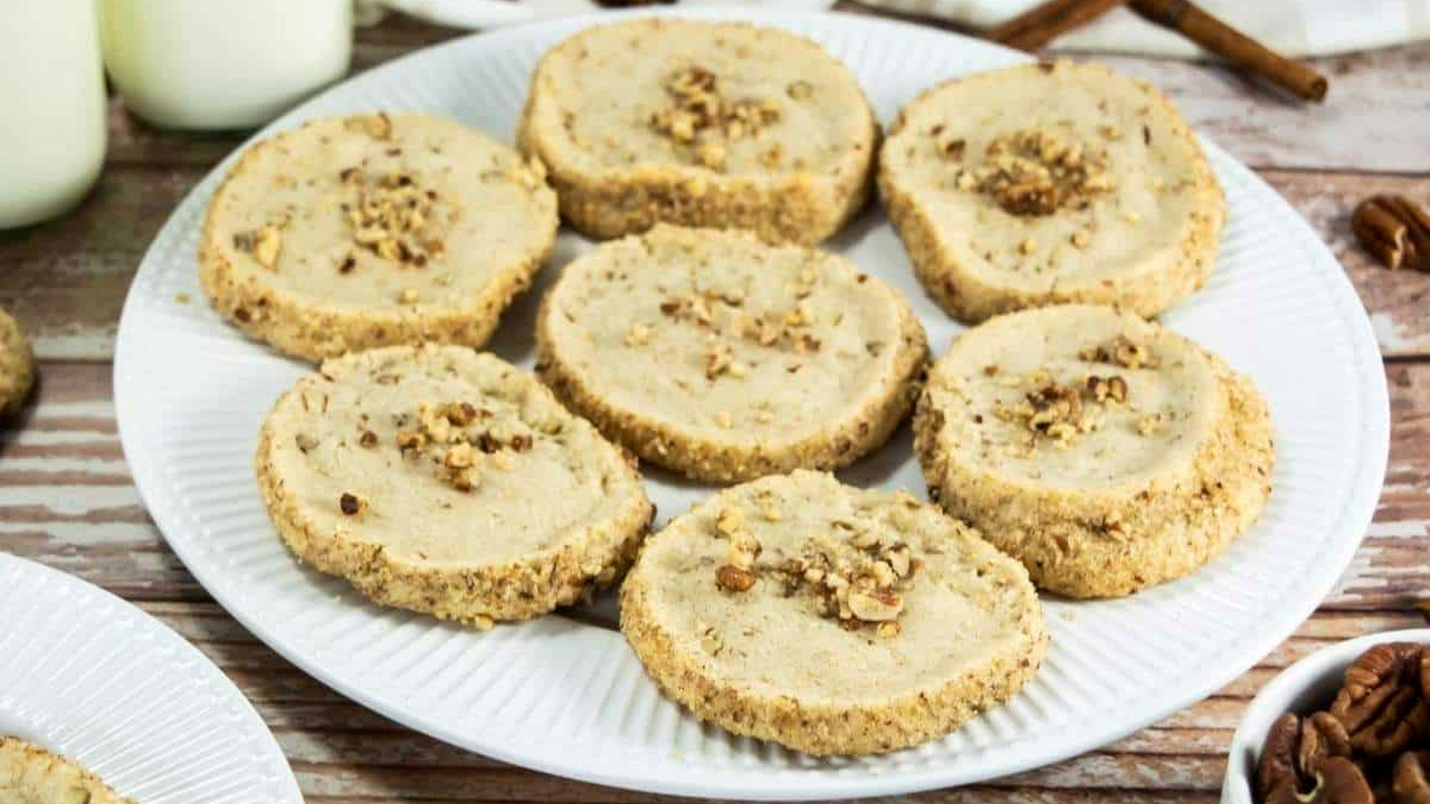 A white plate holds eight round Pecan Shortbread Cookies topped with chopped nuts, placed on a wooden surface next to bottles of milk and cinnamon sticks.