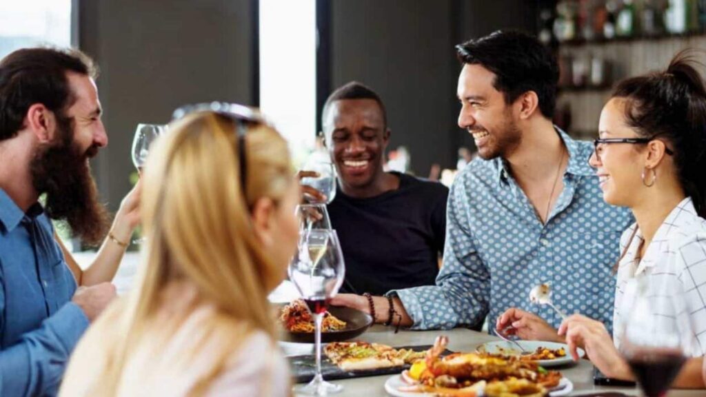 Five people sit around a table, eating food and holding wine glasses, engaged in conversation and smiling in a restaurant setting.
