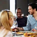 Five people sit around a table, eating food and holding wine glasses, engaged in conversation and smiling in a restaurant setting.
