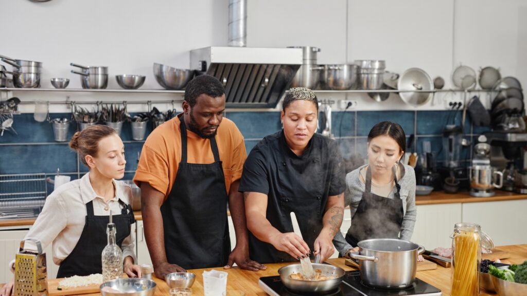 Four people in aprons engage in sight-doing as they watch a chef cook at a kitchen counter, surrounded by pots, pans, and fresh ingredients.