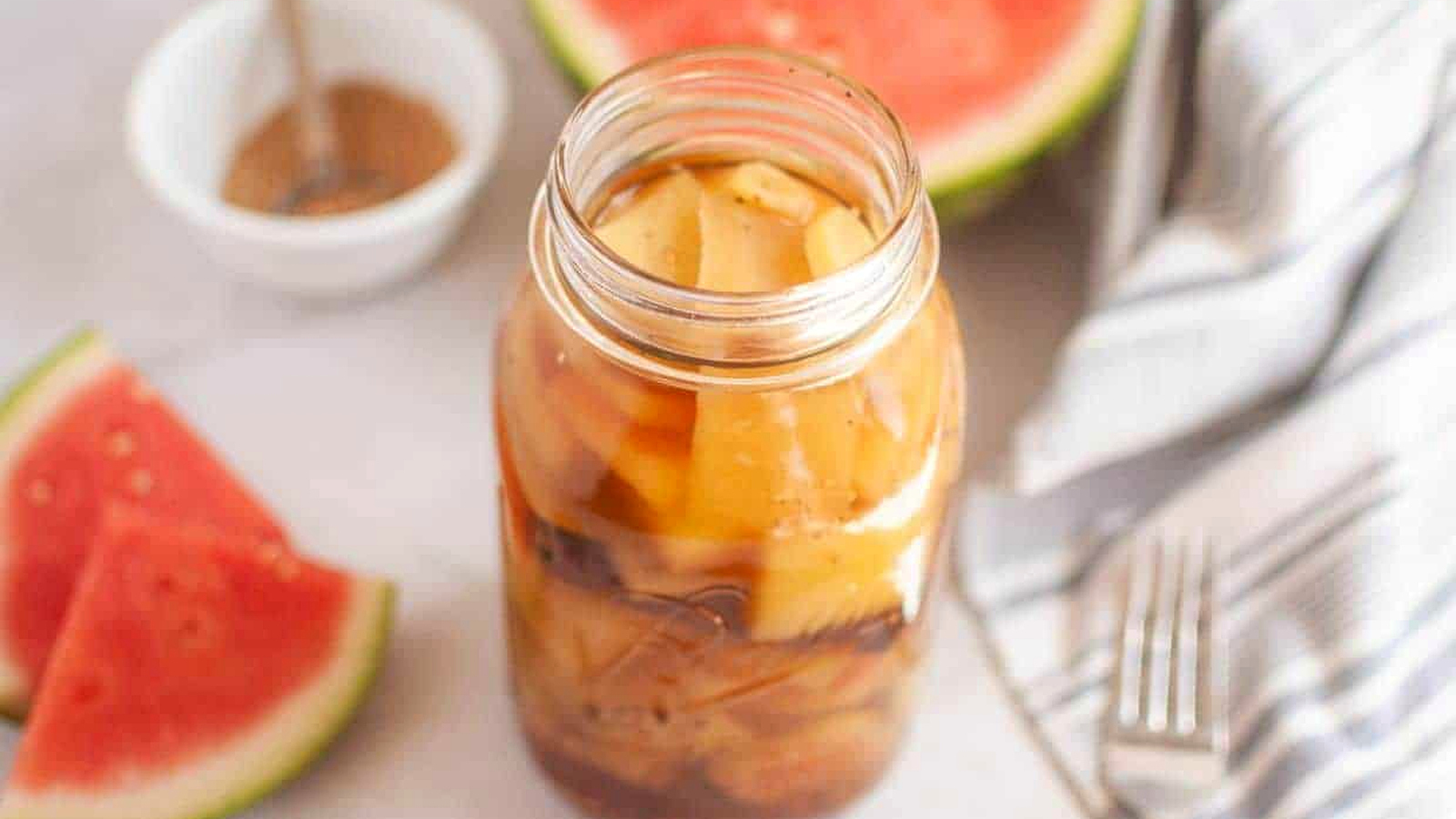 A jar of pickled watermelon rind sits on a white surface, with watermelon slices, a striped cloth, a fork, and a small bowl of spices in the background.
