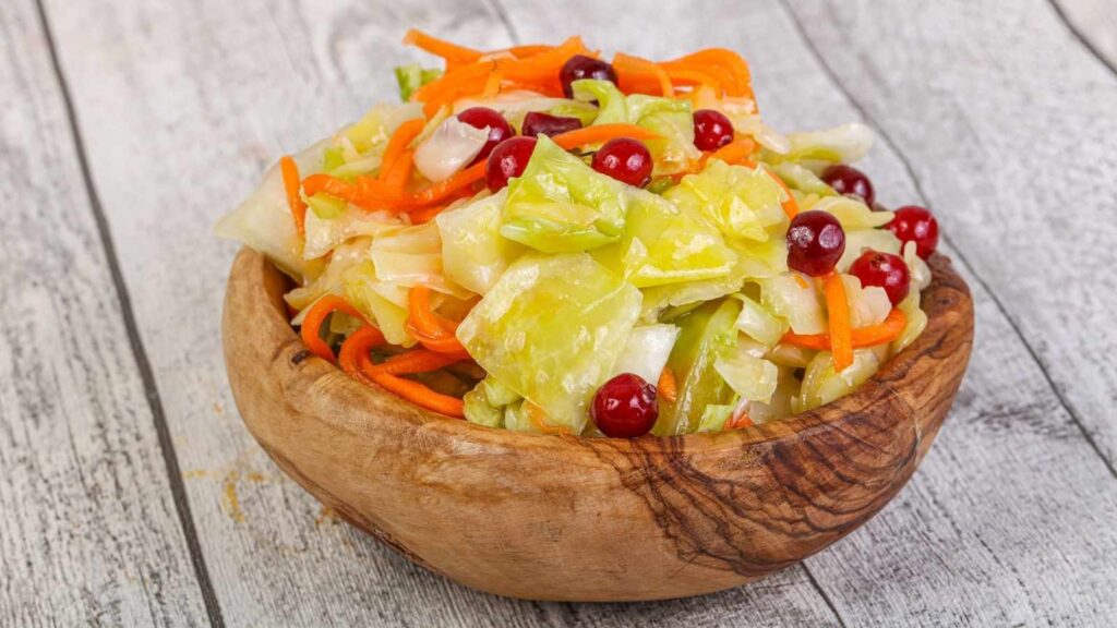 A wooden bowl filled with chopped cabbage, shredded carrots, and red berries on a rustic wooden surface.