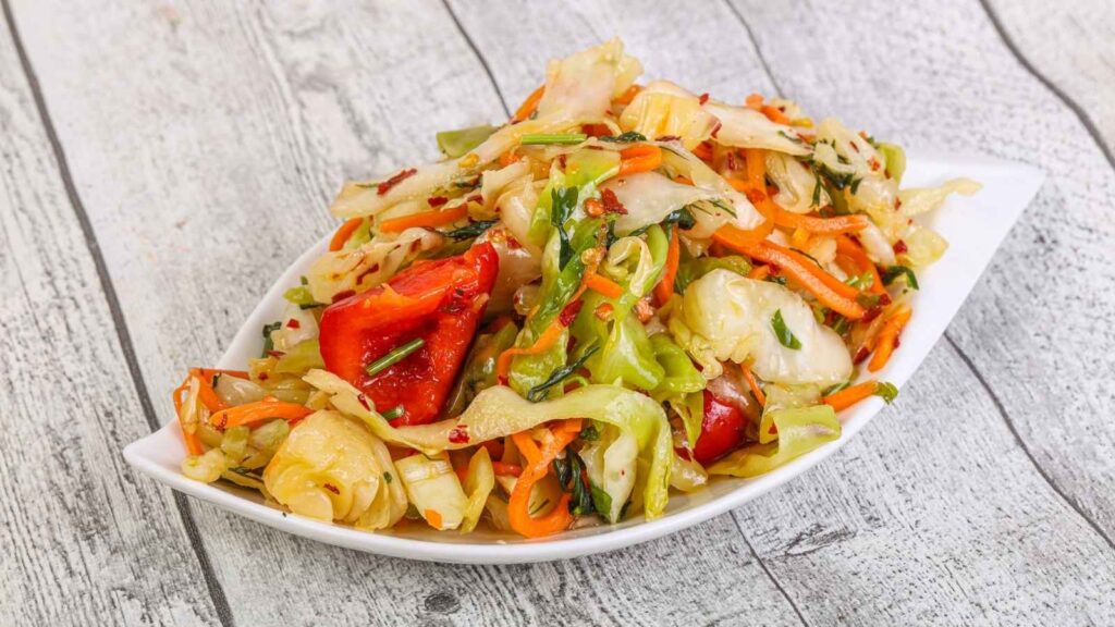 A white dish filled with saut&eacute;ed mixed vegetables, including cabbage, carrots, red bell peppers, and green herbs, showcases a vibrant touch of Haitian food on a wooden surface.