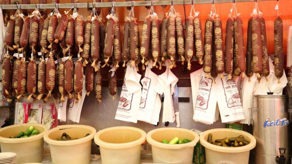 Several cured sausages hang on hooks above white paper bags and large tubs containing pickles in a deli or market setting.