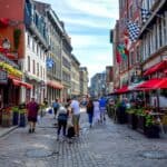 People stroll along a cobblestone street in Québec, lined with shops, cafes, and restaurants featuring outdoor seating and colorful flags—offering a taste of Europe for your next North America vacation.