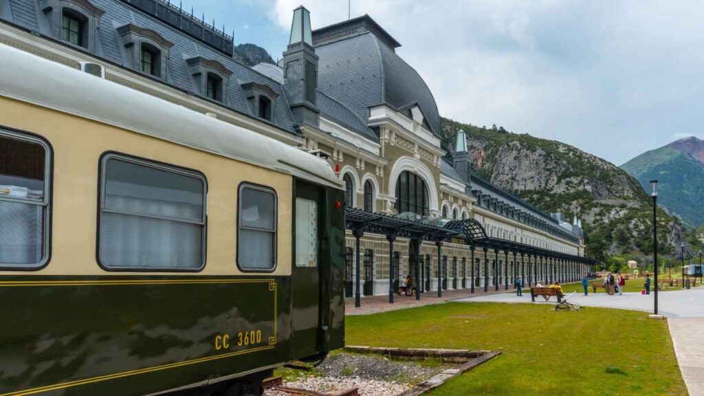 A vintage train is parked beside a large historic railway station with an arched roof; people walk nearby, and mountains are visible in the background.