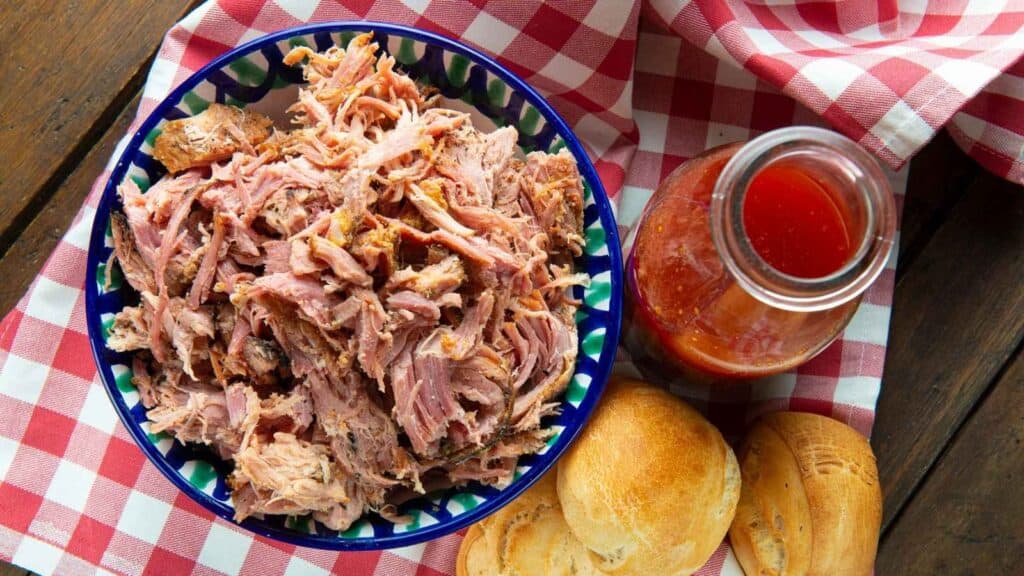 A bowl of shredded pulled pork next to a glass bottle of barbecue sauce and two round bread rolls on a red and white checkered tablecloth.