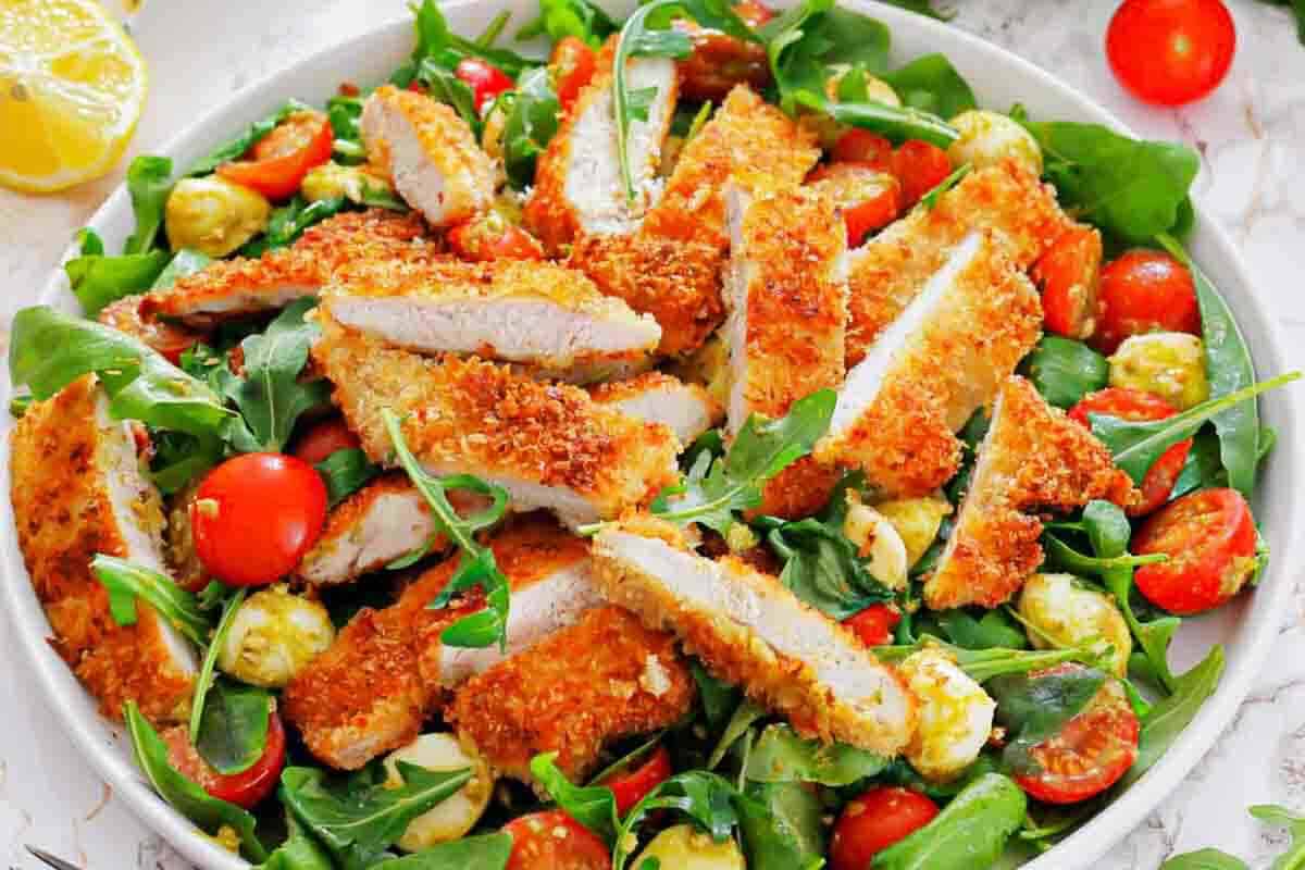 A plate of salad with arugula, cherry tomatoes, mozzarella balls, and sliced breaded chicken cutlets on top.