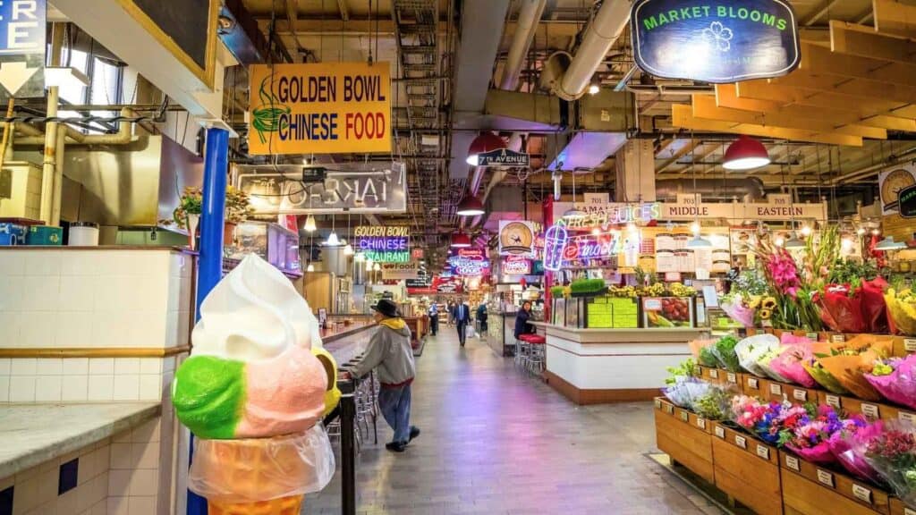 Indoor market with food stalls, neon signs, a large ice cream cone display, and a flower stand on the right. People are walking down the central aisle.