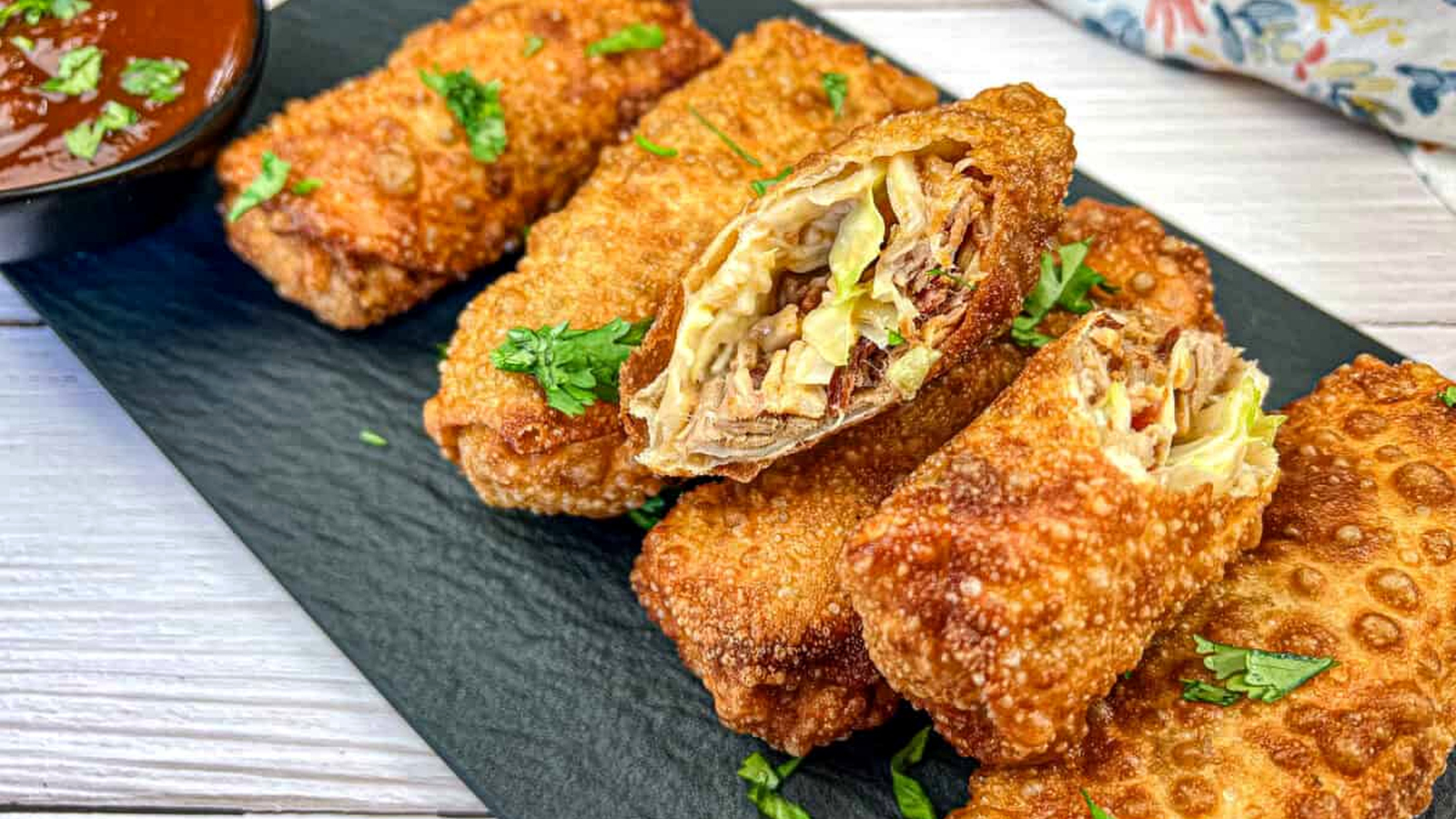 Redneck Eggrolls on a black slate garnished with chopped herbs and a small bowl of dipping sauce.