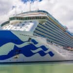 Large white and blue cruise ship docked at a port, with multiple decks and balconies visible on a partly cloudy day.