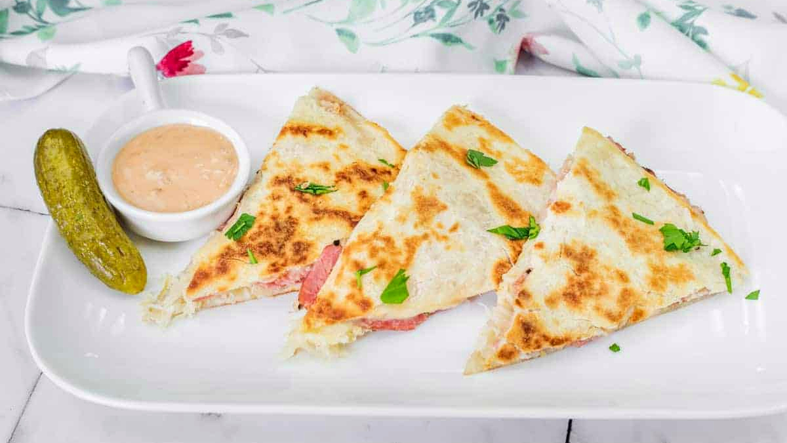 Three Reuben quesadilla slices with herbs on a white plate, accompanied by a pickle and a small dish of dipping sauce.