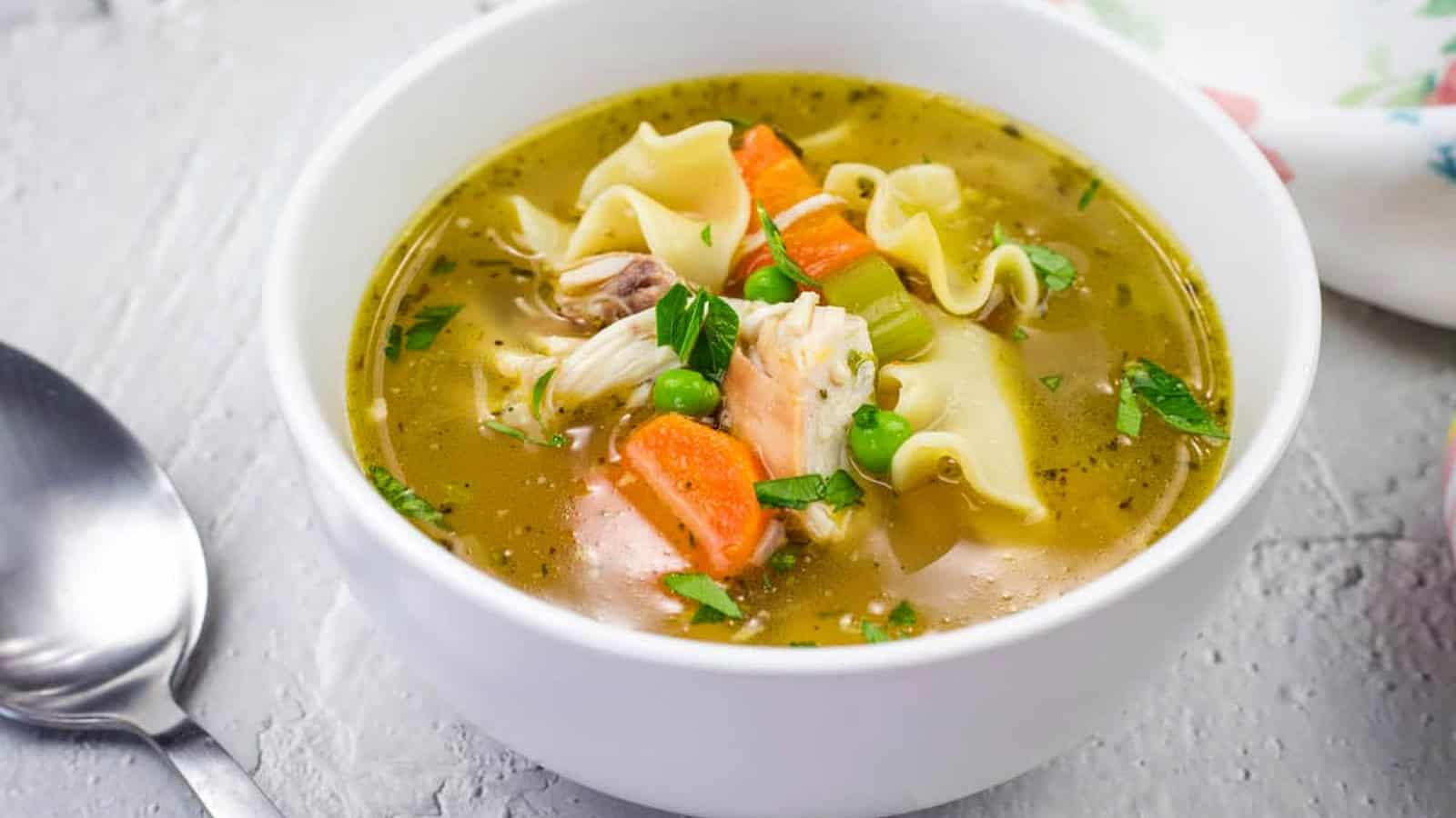 A bowl of Rotisserie Chicken Noodle Soup topped with parsley on a white surface, next to a spoon.