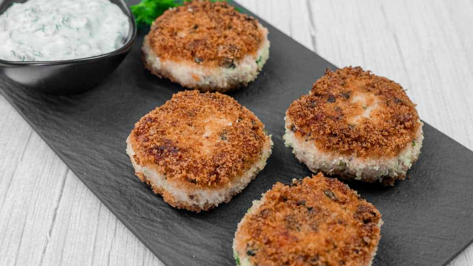 Four Salmon Cakes are arranged on a black slate serving board, accompanied by a small bowl of white sauce with herbs.