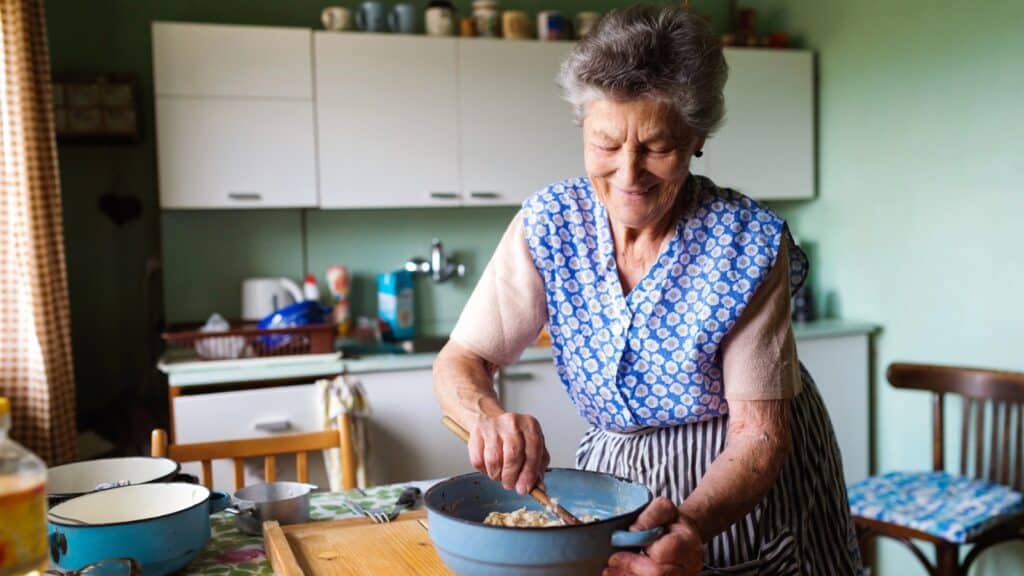An elderly woman wearing an apron stands in a kitchen, stirring a mixture in a bowl on a table covered with a floral tablecloth.