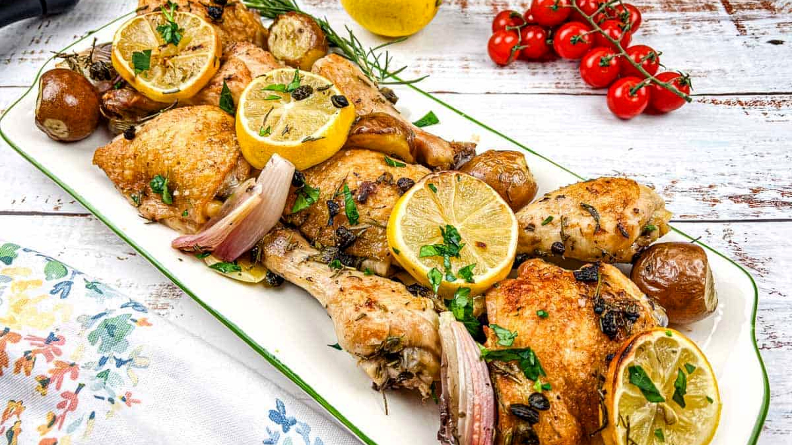 A platter of sheet pan lemon-rosemary chicken with lemon slices, herbs, and garnished with cherry tomatoes.