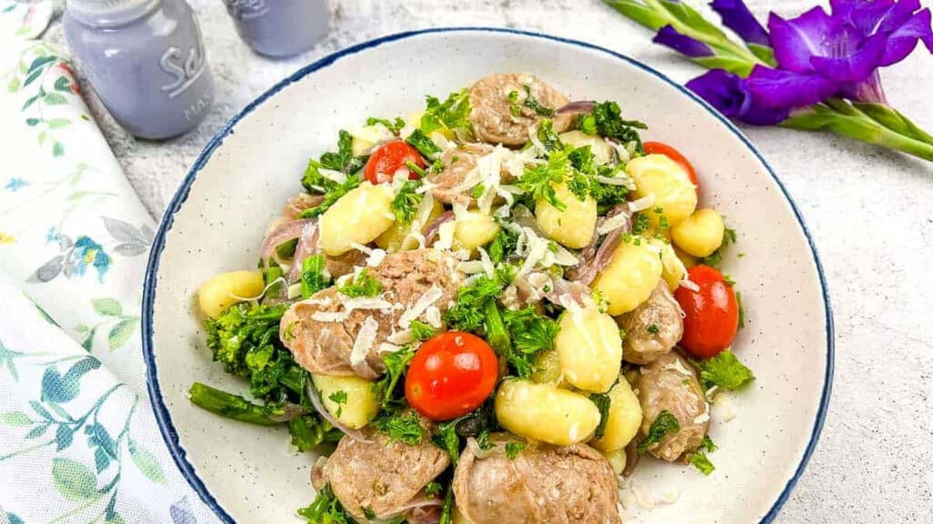 A bowl of gnocchi with sausage, broccoli, cherry tomatoes, grated cheese, and herbs, placed on a light surface beside purple flowers and salt and pepper shakers.