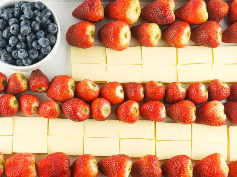 A tray arranged to resemble the U.S. flag with blueberries in a bowl representing the stars, and alternating rows of cheese slices and strawberries representing the stripes.