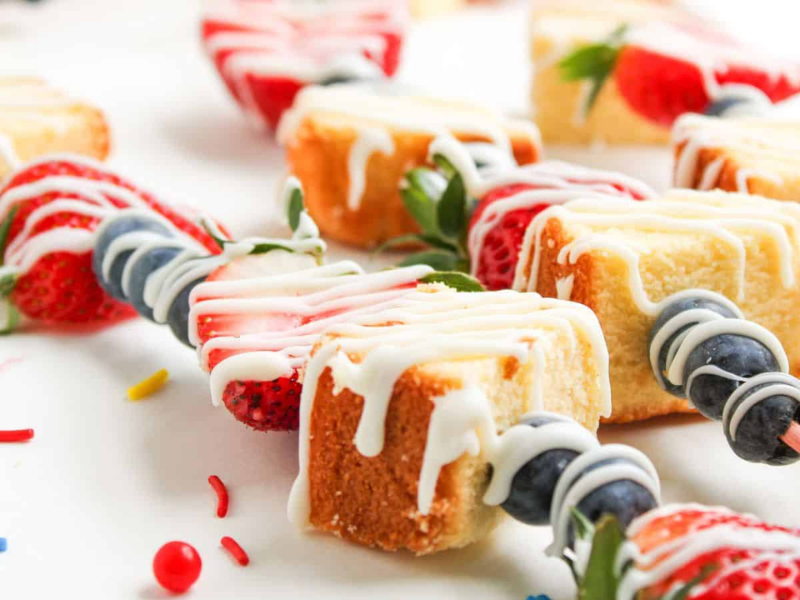 Strawberry, blueberry, and pound cake pieces on skewers, drizzled with white icing, arranged on a white surface with colorful sprinkles.