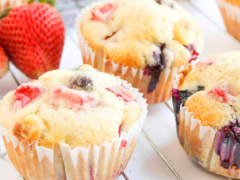 Three berry muffins with visible strawberry and blueberry pieces sit on a cooling rack next to whole strawberries.