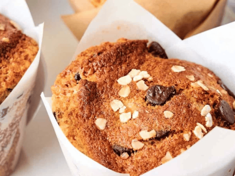 A close-up of a muffin topped with oats and chocolate chips, wrapped in white parchment paper.