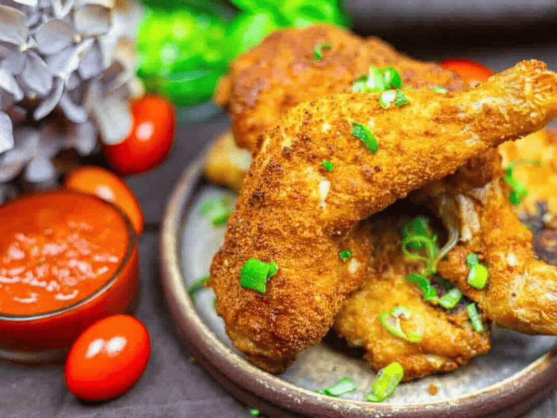 A plate of crispy fried chicken garnished with chopped green onions, served with a side of red dipping sauce and cherry tomatoes.
