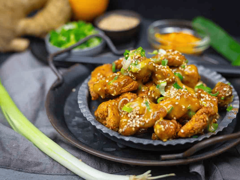 A plate of sesame-coated, sauced chicken pieces garnished with sliced green onions. Various fresh ingredients and spices are visible in the background.