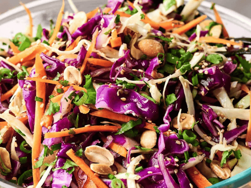 A bowl of colorful coleslaw with shredded purple and green cabbage, carrots, chopped peanuts, and greens, mixed with dressing.