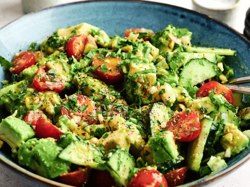 A blue bowl filled with a fresh salad containing avocado, cucumber, cherry tomatoes, herbs, and a light dressing, with a fork resting on the side.