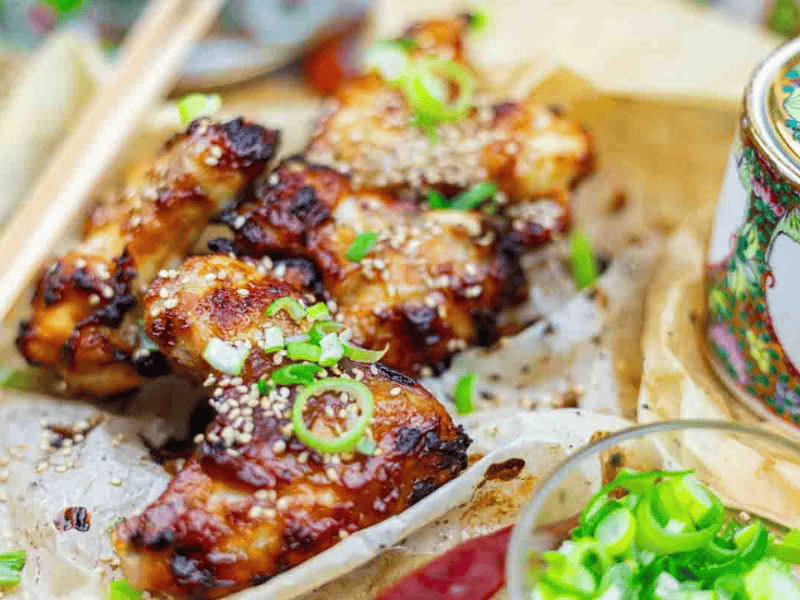 BBQ chicken wings topped with sesame seeds and chopped green onions, served on parchment paper with a side dish partially visible.