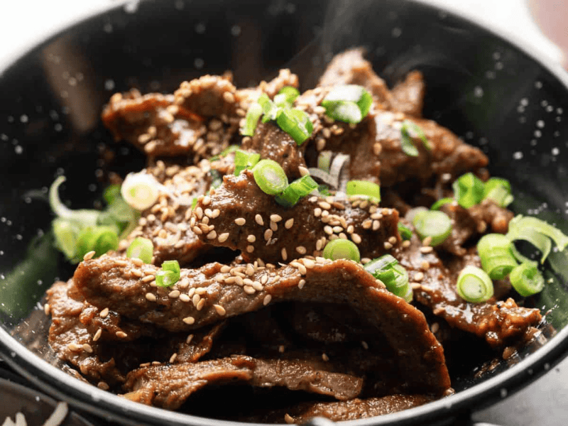 A bowl of cooked beef slices topped with sesame seeds and chopped green onions.