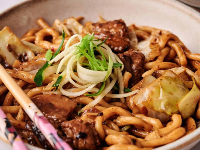 A bowl of stir-fried udon noodles with beef, cabbage, and green onion garnish, served with a pair of chopsticks on the side.