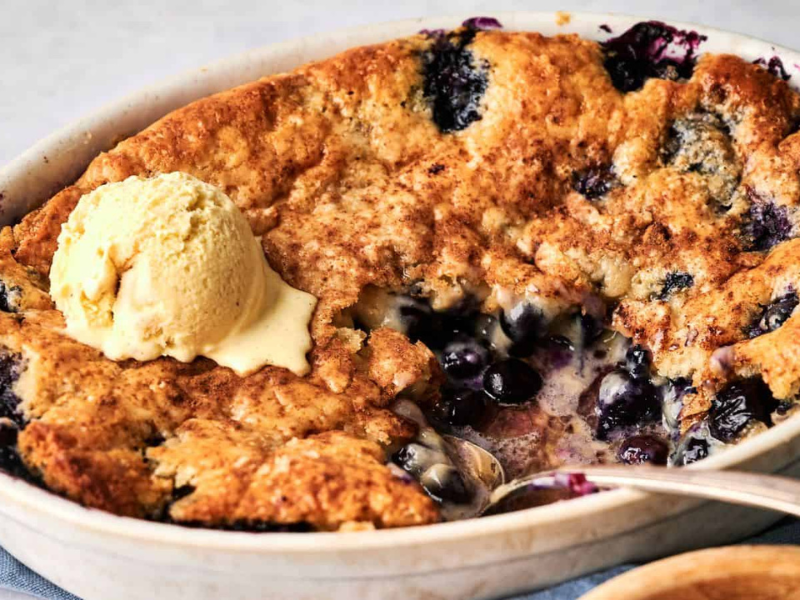 A baked blueberry cobbler with a golden crust is shown in a white dish, partially served, topped with a scoop of vanilla ice cream.