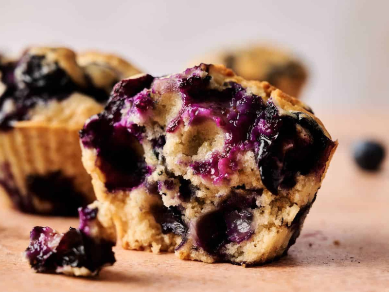 A close-up of a blueberry muffin with a bite taken out, showing the moist interior and whole blueberries; other muffins and loose blueberries are in the background.
