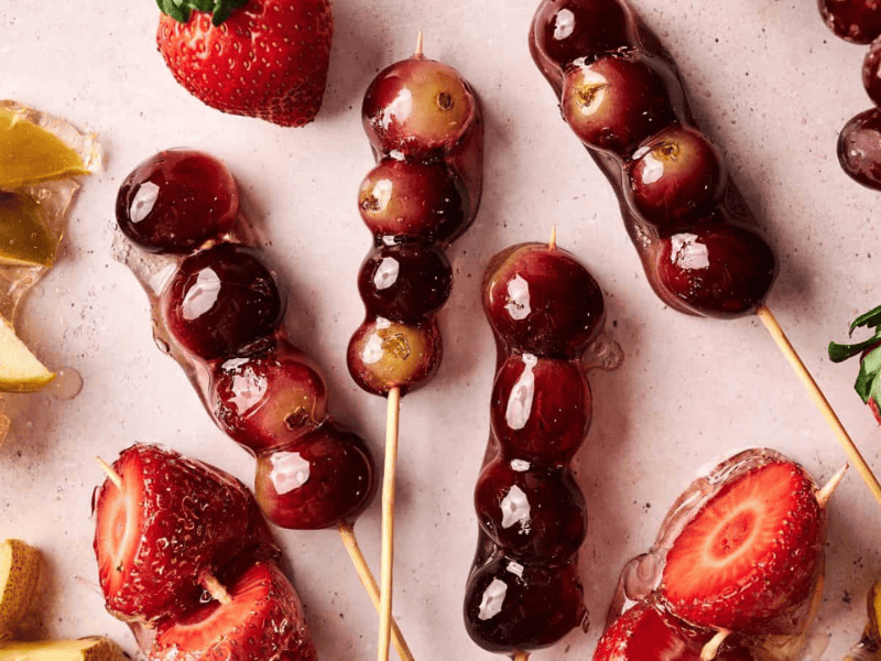 Skewers of grapes and strawberries coated in a shiny glaze are arranged on a light surface with loose fruit pieces nearby.