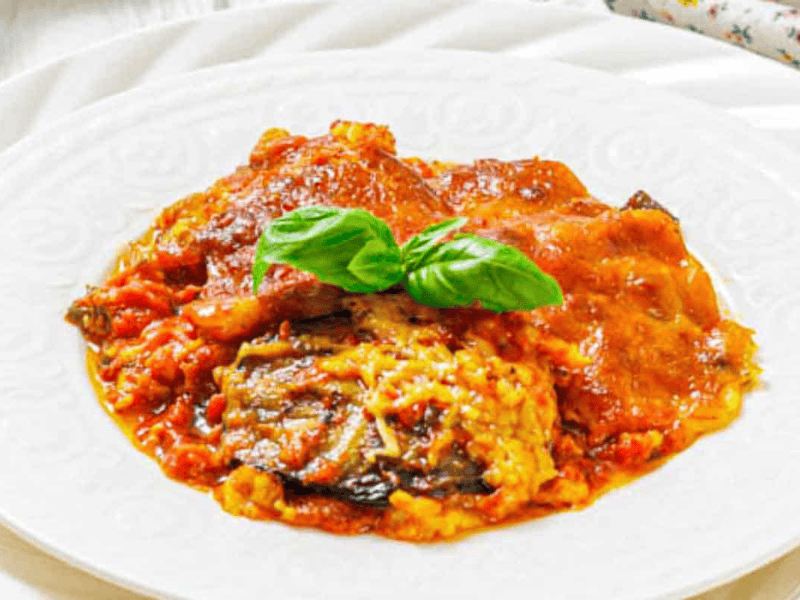 A serving of eggplant parmesan topped with tomato sauce and melted cheese, garnished with fresh basil leaves on a white plate.