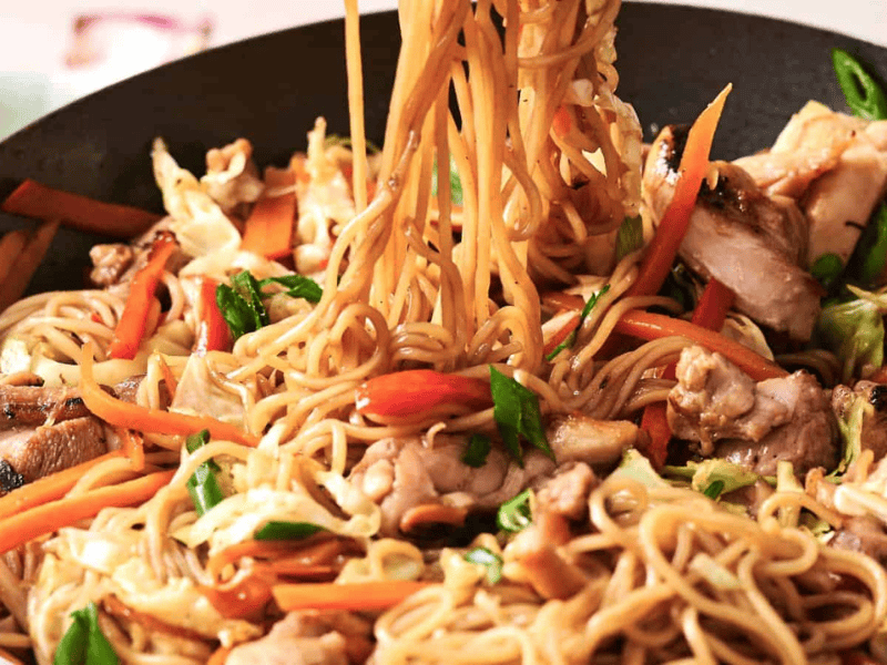 Stir-fried noodles with vegetables and chicken in a pan, featuring carrots, cabbage, and green onions.