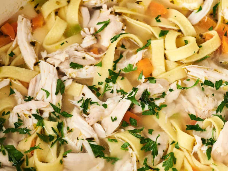 A close-up of a bowl of hearty Chicken Noodle Soup, showcasing shredded chicken, egg noodles, chopped carrots, celery, and garnished with fresh parsley.