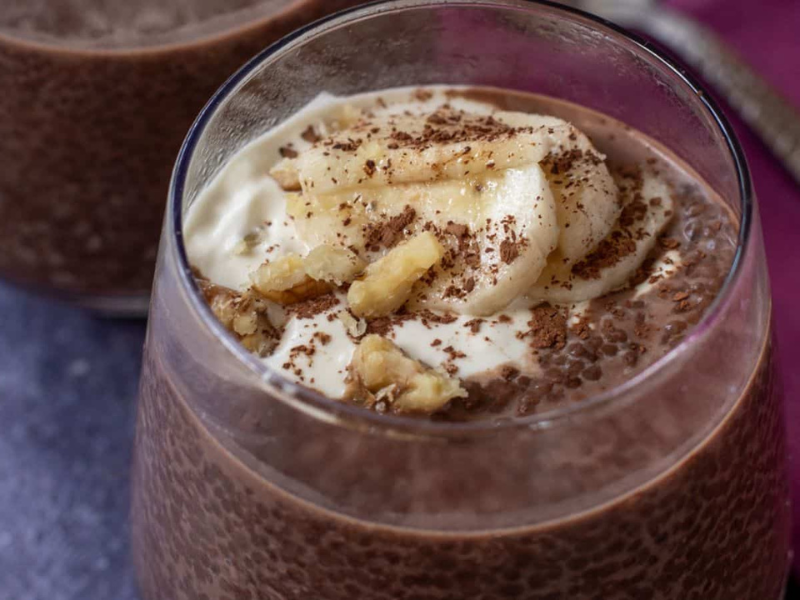 A glass of chocolate chia pudding topped with banana slices, yogurt, chopped walnuts, and chocolate shavings, with another glass partially visible behind it.