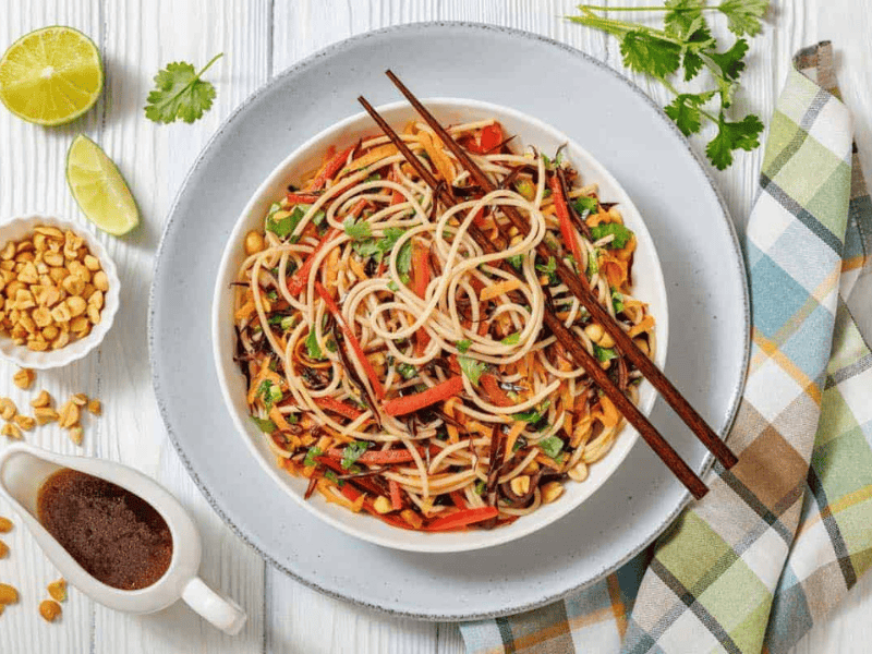 A bowl of Asian noodle salad with vegetables and chopsticks, surrounded by lime, peanuts, sauce, and fresh cilantro on a light wooden table with a plaid napkin.