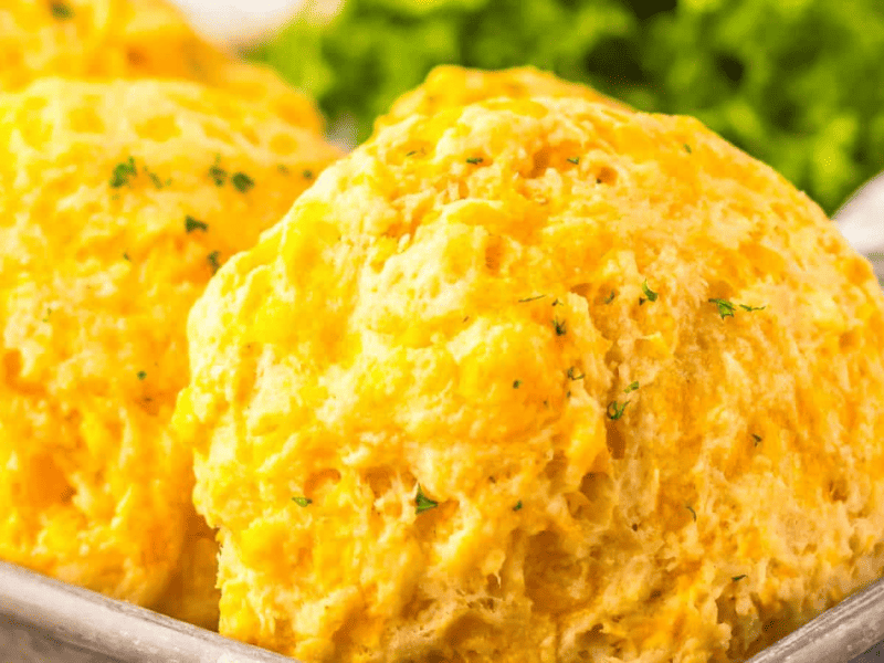 Close-up of fluffy cheddar biscuits topped with herbs on a metal tray, with leafy greens in the background.