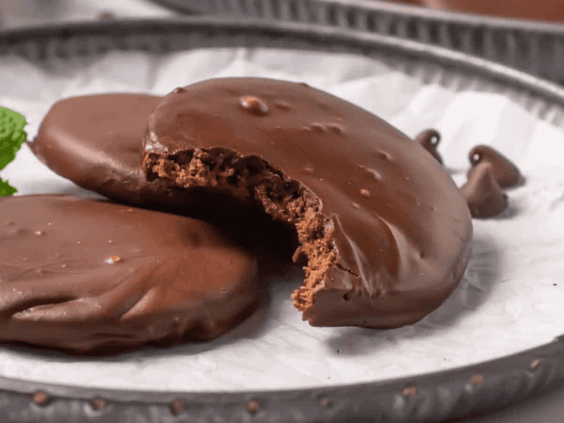 Two copycat thin mint cookies on a plate, one with a bite taken out, with a mint leaf garnish beside them.