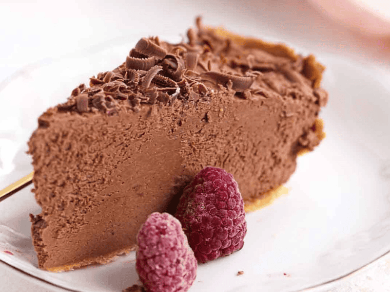 A slice of chocolate mousse pie on a white plate, topped with chocolate shavings and served with two raspberries.