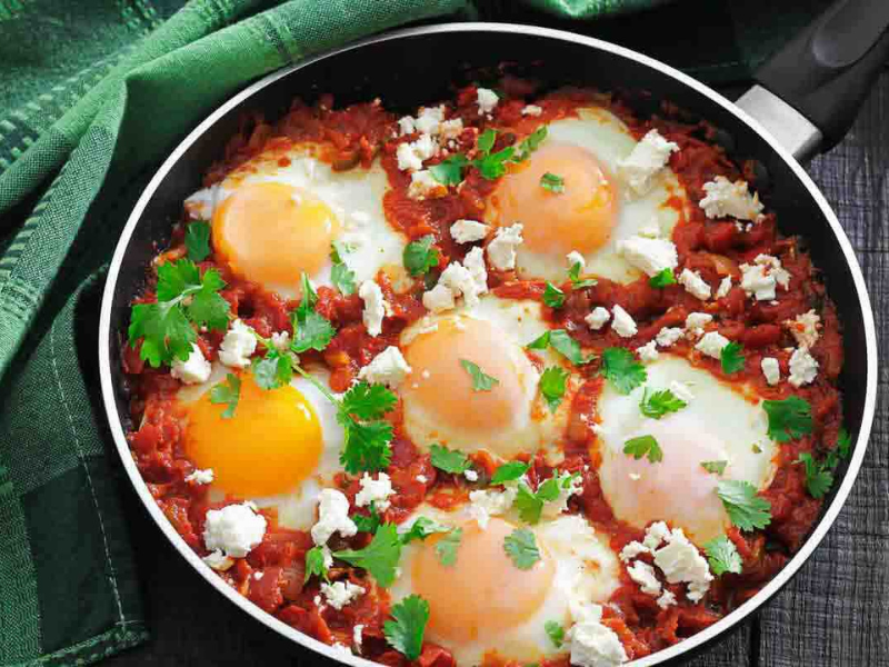 A pan of shakshuka with poached eggs in tomato sauce, garnished with fresh herbs and crumbled cheese, sits on a dark table next to a green cloth.